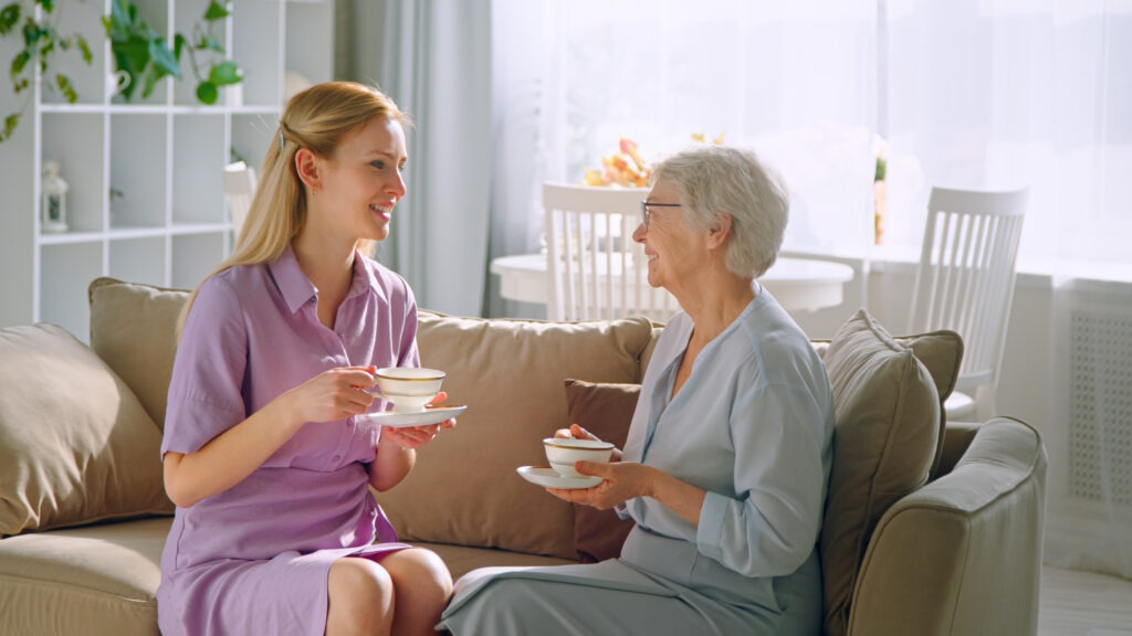 Cuidadora e idosa em conversa descontraída a tomar chá, sorriso genuíno, ambiente de sala com luz natural.