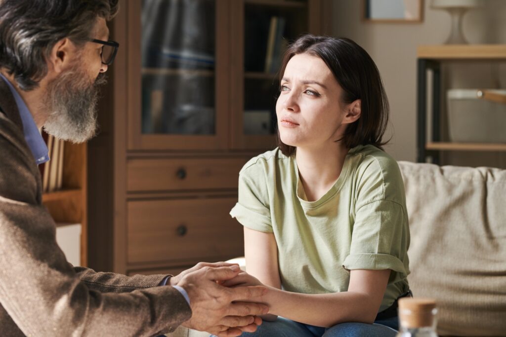 Conversa séria entre duas pessoas em casa, com contacto físico nas mãos, expressão atenta e preocupada, mas não dramática.
