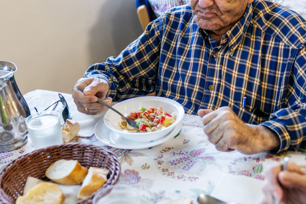 Homem idoso a comer uma refeição à mesa, postura concentrada, ambiente de cuidado e rotina.